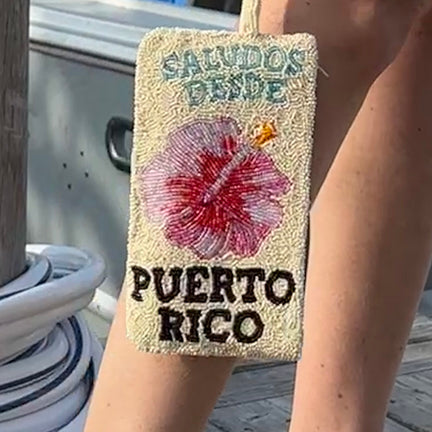 Beaded purse with pink flower and text 'Saludos desde Puerto Rico' held by a person. With "Saludos Desde Puerto Rico" stitched in bold, island-inspired lettering and a vibrant hibiscus in full bloom, this piece is a love letter to tropical days and unforgettable getaways.